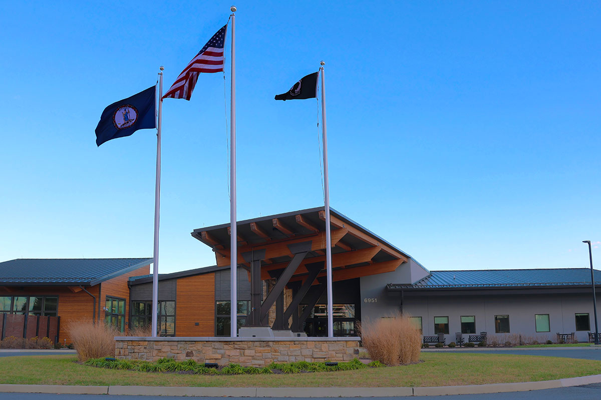 Exterior view of the Puller Veterans Care Center, showing the main entrance with three flagpoles flying the U.S, flag, the Virginia state flag, and the POW/MIA flag in front of the building under a clear blue sky.
